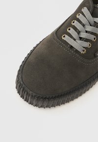 Baskets en daim gris avec une semelle en caoutchouc noir texturée, des lacets gris et des accents dorés aux œillets. Surface lisse avec des détails de couture visibles.