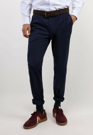 Hombre con pantalones azul marino remangados en los tobillos, calcetines verdes, zapatillas burdeos con rayas naranjas y blancas, cinturón marrón y camisa a rayas azul claro.