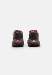 Brune og maroon sneakers med polstrede krager, teksturerte såler og merkede hælfliker. Har logodetaljer på siden og røde aksenter.
