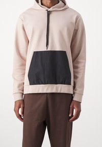 Sudadera de color beige claro con bolsillo de tela negra, capucha con cordón y dobladillo elástico. Material suave con un corte relajado. Diseño minimalista.