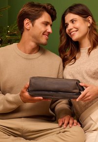 Hombre y mujer sonriendo el uno al otro mientras sostienen un pouch de cuero oscuro, sentados frente a un árbol de Navidad decorado con un fondo verde.
