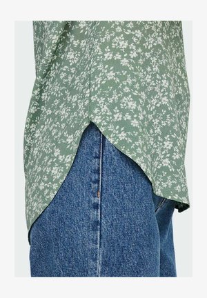 Zöld virágmintás felső, ívelt díszítéssel, kék farmernadrággal párosítva, amely középen gombbal és cipzárral záródik.