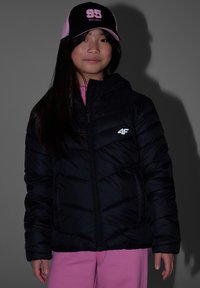 Fille portant une doudoune noire, un pantalon rose et une casquette de baseball rose et noire, debout devant un fond gris.