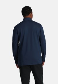 Marineblaues langärmliges Polohemd mit Kragen, das eine glatte Textur und dezente Markenkennzeichnung auf dem oberen Rücken aufweist. Getragen mit schwarzen Hosen.