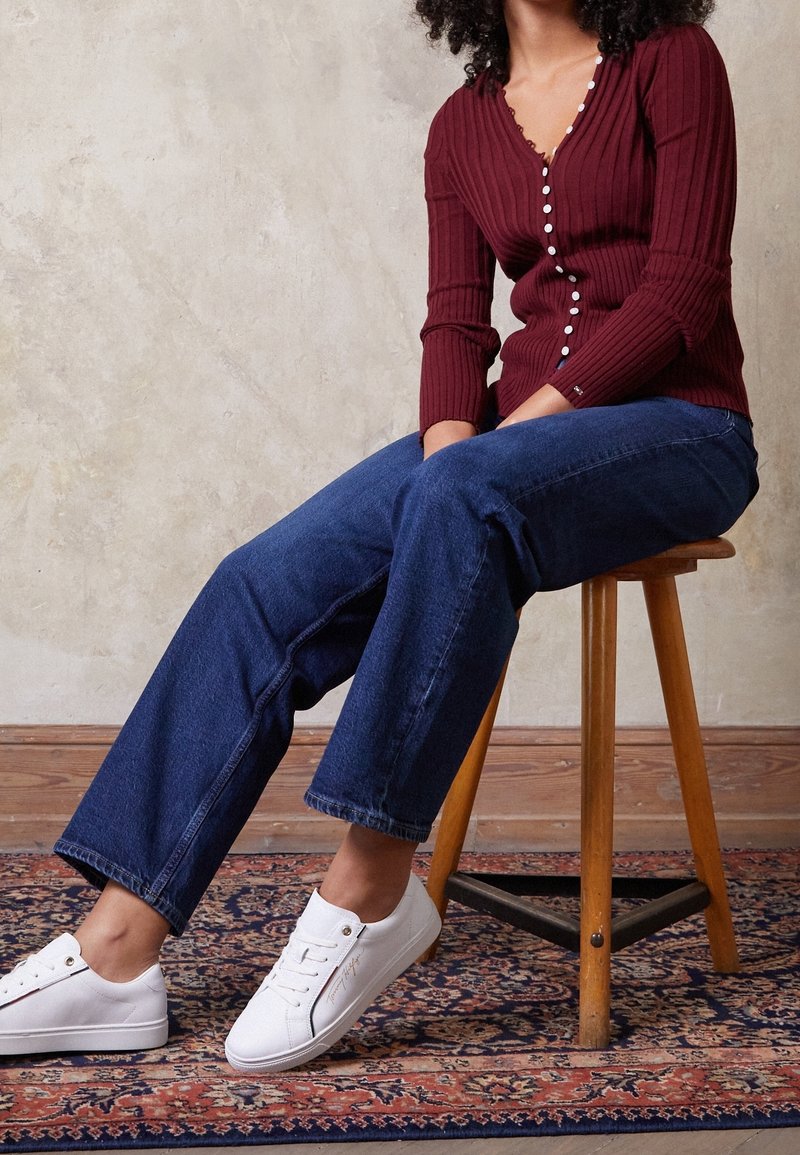 Cardigan a coste bordeaux con bottoni, abbinato a jeans in denim blu a gamba larga e sneakers bianche, seduto su uno sgabello di legno.