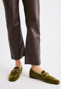 Pies con mocasines de terciopelo verde oliva y acentos de hebilla metálica dorada, combinados con pantalones de cuero marrón oscuro recortados sobre fondo blanco.