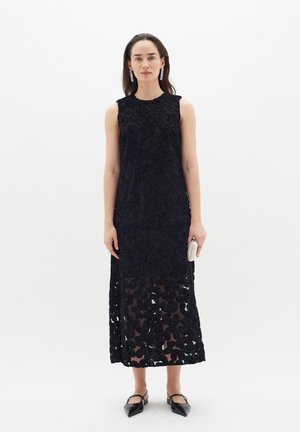 Schwarzes Spitzenkleid mit floralem Muster, ärmelloses Design und durchsichtiger Rock. Die Modell hält eine kleine weiße Clutch und trägt spitze schwarze Schuhe.