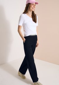 Jonge vrouw lacht, draagt een roze pet, wit T-shirt met korte mouwen, zwarte broek met zakken en beige sneakers, loopt binnen.