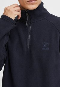 Mörk marinblå fleecepullover med hög krage, halvdragkedja fram och en broderad logotyp på bröstet. Texturerad tyg med en mjuk känsla.
