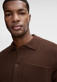 Brązowa dziana koszula z kołnierzykiem, z przednią kieszenią i zapięciem na dwa guziki. Teksturowana tkanina w jednolitym kolorze i prostym wzorze.