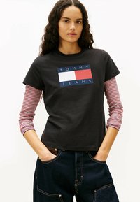 Czarna bawełniana koszulka z niebieskim, białym i czerwonym prostokątnym logo z napisem "TOMMY JEANS." Długie rękawy w czerwono-białe paski widoczne spod spodu.