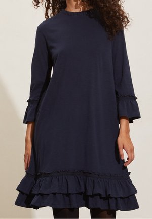 Vrouw in een marineblauwe jurk met een geraffineerde rand en manchetten, staande tegen een lichte beige achtergrond.