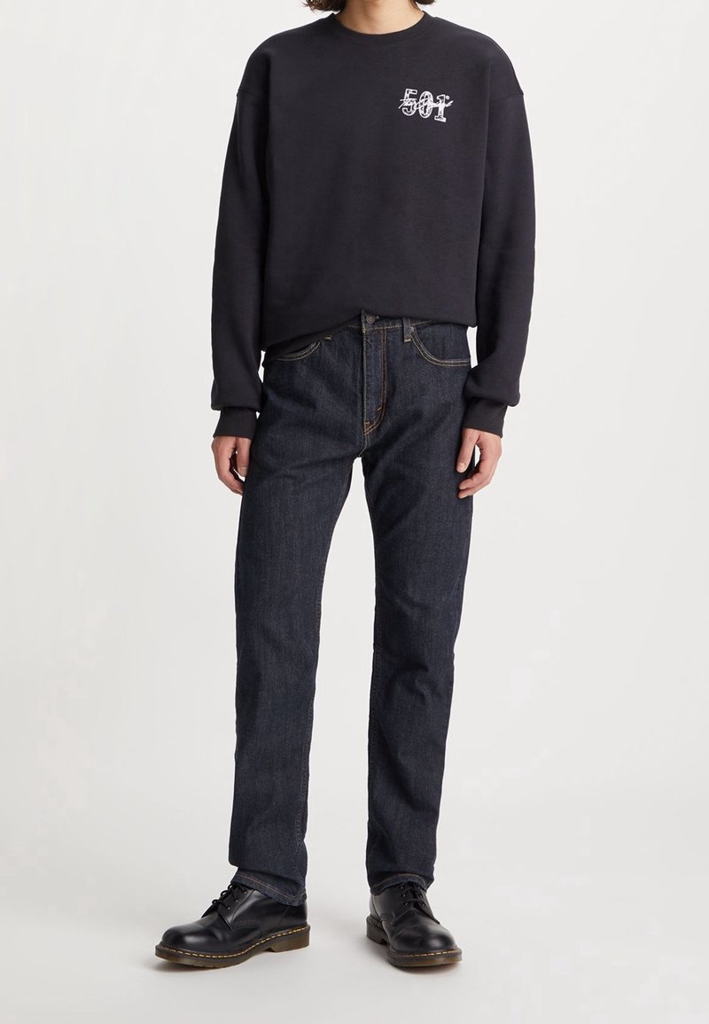 Personne portant une chemise noire à manches longues avec le texte "501", un jean en denim bleu foncé et des chaussures noires à lacets, debout devant un fond uni.