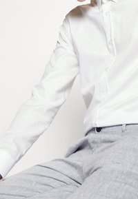 Person wearing a white long-sleeve buttoned shirt and light gray pants, sitting against a plain white background.