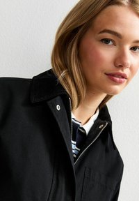 Svart jacka med krage och dragkedja, med en framficka och textilstruktur. Under jackan syns en vit och marinrandig skjorta.