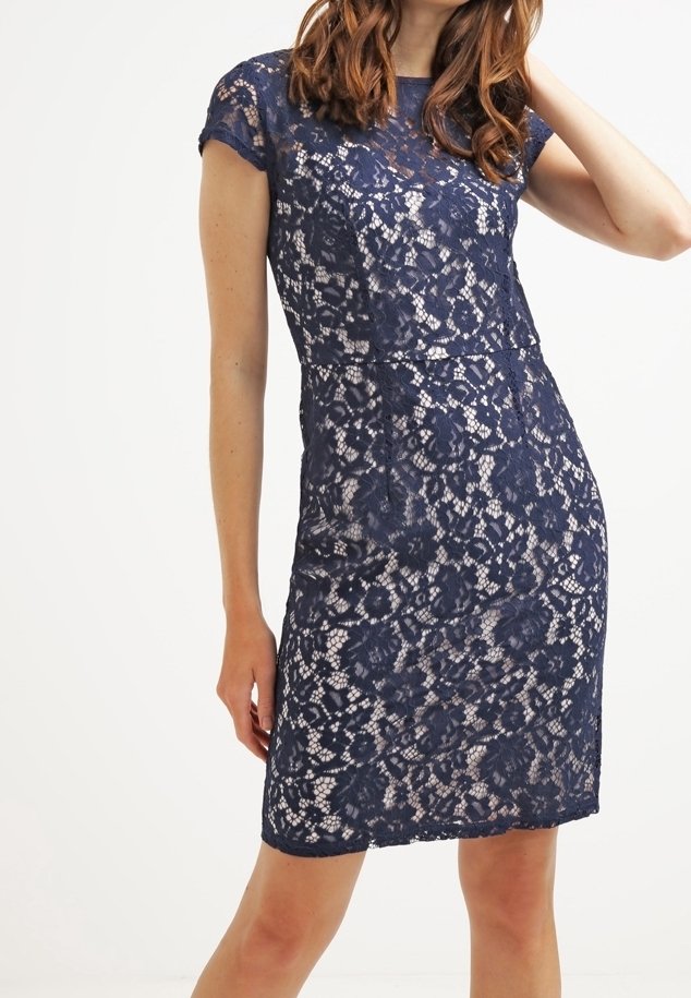Námorně modré květinové krajkové šaty s krátkými rukávy, které nosí žena, s přiléhavým střihem končícím nad koleny na bílém pozadí.