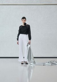 Blusa nera trasparente con maniche a balze, pantaloni bianchi a palazzo e tacchi neri con cinturini, mentre tiene un tessuto argentato lucido drappeggiato.