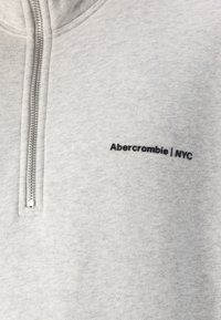 Sudadera gris con cremallera hecha de tela suave; presenta un logo negro "Abercrombie | NYC" en el lado izquierdo del pecho y una cremallera metálica.
