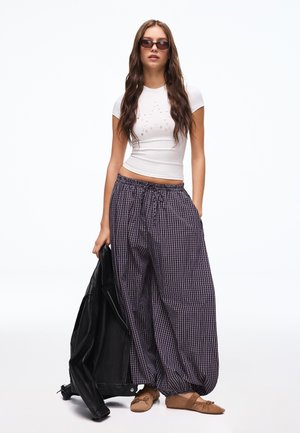 Femme aux longs cheveux bruns portant des lunettes de soleil, un top blanc ajusté à manches courtes, un pantalon ample à carreaux, des ballerines marron, tenant une veste noire.