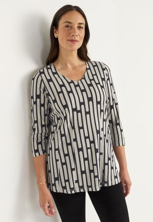 Vrouw leunt tegen een muur en draagt een grijs en zwart geometrisch patroon V-hals top met driekwart mouwen en zwarte broek.