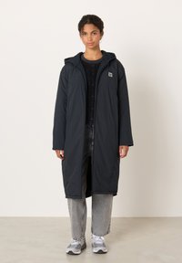 Ung kvinna klädd i en lång svart huva, svart tröja, grå jeans och vita-grå sneakers som står mot en enfärgad bakgrund.