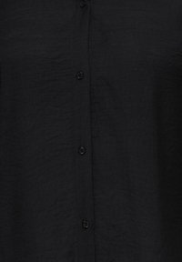 Svart skjorte i teksturert stoff med en vertikal rad av fem små matchende svarte knapper nedover midten.