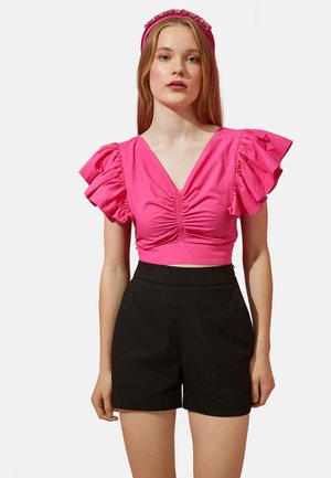 Roze crop top met v-hals en volants mouwen. Heeft gerimpelde details aan de voorkant. Gecombineerd met een hoge taille zwarte shorts.