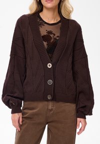 Brązowy dzianinowy kardigan z dekoltem w serek, wzorami teksturowymi i trzema dużymi guzikami. Posiada szerokie rękawy i skrócony dół.