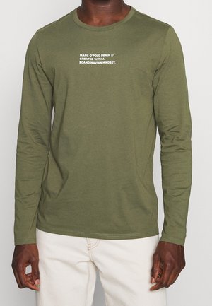 Man draagt een olijfgroen shirt met lange mouwen en witte tekst "Marc O'Polo Denim gemaakt met een Scandinavische denkwijze" en lichtgekleurde broek.