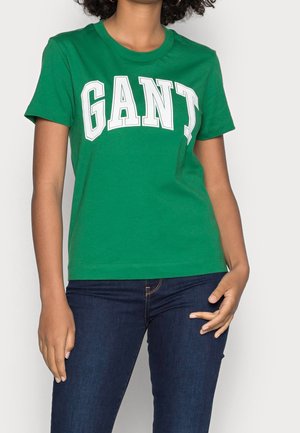 Osoba ubrana w zielony T-shirt z krótkim rękawem z białym napisem "GANT" i ciemnoniebieskie dżinsy, stojąca na jednolitym jasnym tle.