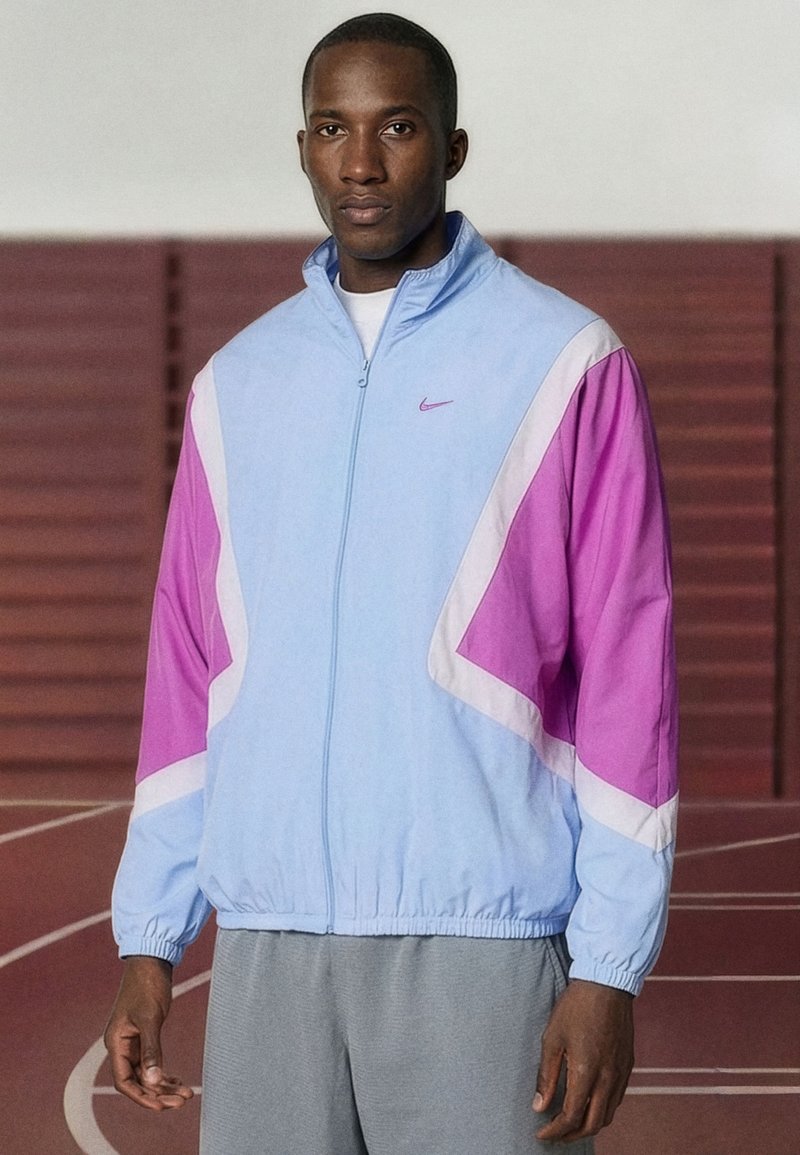 Man staat op een indoorbaan, draagt een lichtblauw en paars sportjack met ritssluiting en een grijze broek, kijkt naar voren met een neutrale uitdrukking.