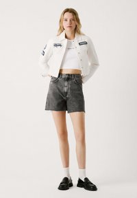 Chaqueta de mezclilla blanca con parches de logo, combinada con pantalones cortos de mezclilla negra de tiro alto. Mocasines negros y calcetines blancos completan el look.
