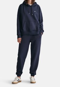 Persona vestita con una felpa blu navy e pantaloni da jogging coordinati, abbinati a sneakers bianche e blu navy, in piedi contro uno sfondo neutro.