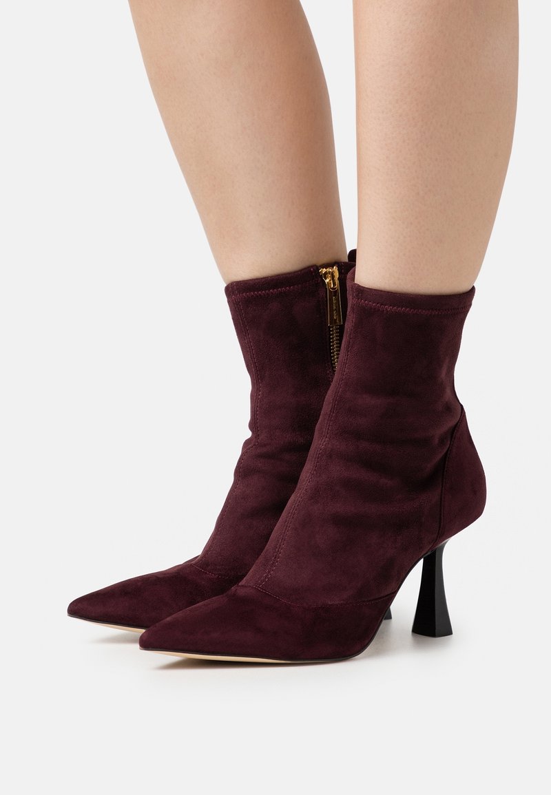 Paire de bottines en daim bordeaux foncé avec bouts pointus, fermetures éclair latérales dorées et talons carrés évasés portées sur jambes nues.