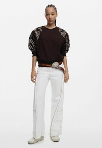 Sweatshirt maro cu umeri embellisheti, blugi albi cu picioare late și cusături în contrast, și o curea maro cu o cataramă argintie.