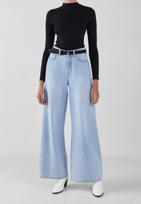 Jean large bleu clair en denim, avec un haut noir à manches longues côtelé et une ceinture noire cloutée. La tenue est complétée par des talons blancs.