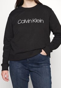 Černá mikina s kulatým výstřihem, se znakem "Calvin Klein" v bílé barvě. Vyrobena z měkké látky, s žebrovanými manžetami a spodním lemem.