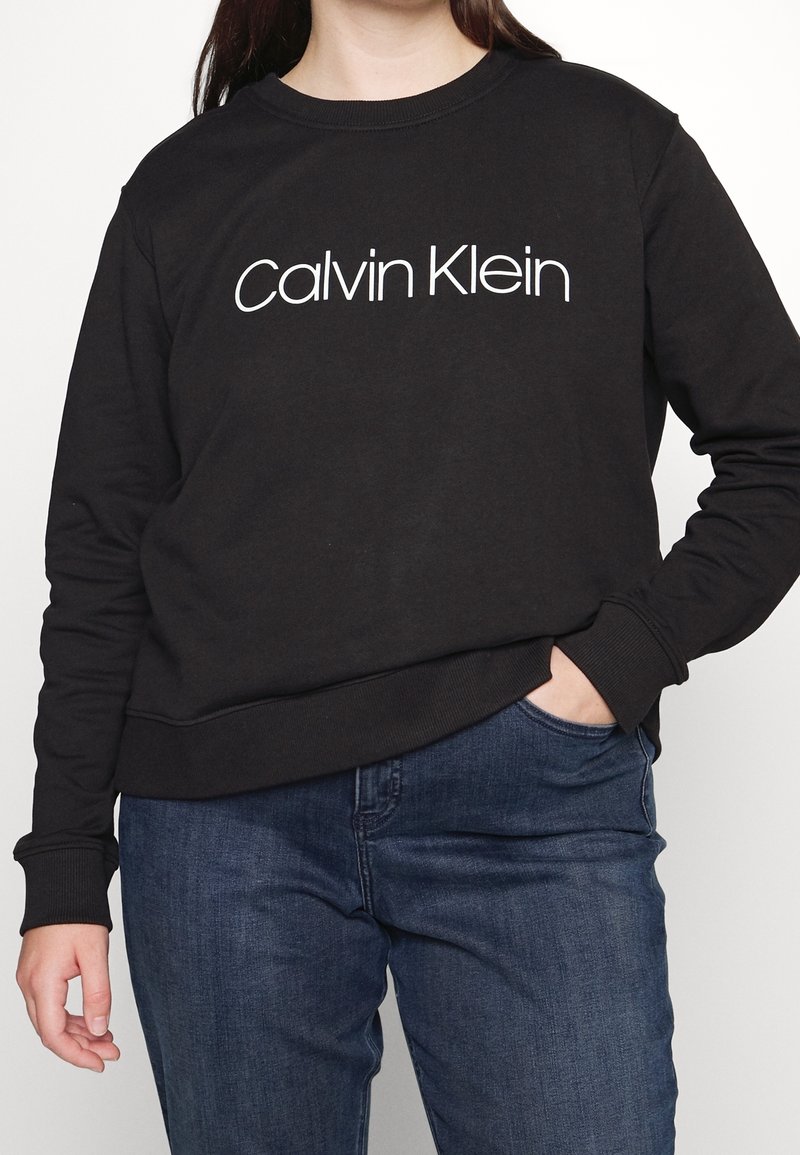 Černá mikina s kulatým výstřihem, se znakem "Calvin Klein" v bílé barvě. Vyrobena z měkké látky, s žebrovanými manžetami a spodním lemem.
