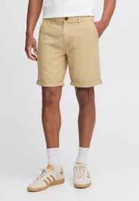 Beige Baumwollshorts mit umgeschlagenen Säumen, mit einem Knopfverschluss und Seitentaschen, kombiniert mit weißen und beigen Sneakern sowie weißen Socken.