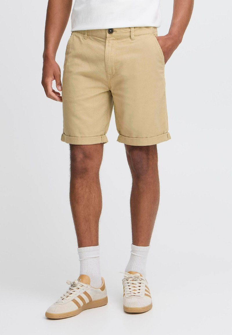 Beige Baumwollshorts mit umgeschlagenen Säumen, mit einem Knopfverschluss und Seitentaschen, kombiniert mit weißen und beigen Sneakern sowie weißen Socken.
