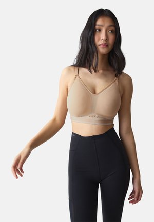 Ung kvinde med langt mørkt hår iført en beige sports-bh og sorte højtaljede leggings mod en ensartet lys baggrund.