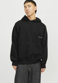 Hoodie en coton noir avec une poche kangourou à l'avant, une capuche spacieuse et un détail textuel discret sur la manche gauche. Conçu avec une coupe décontractée.