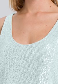 Débardeur en sequins bleu clair avec un décolleté bateau et un tissu texturé. La surface est ornée de sequins réfléchissants agencés en motif.
