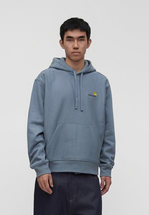 Ung mand iført en blå Carhartt-hættetrøje og mørke bukser, stående foran en ensfarvet lysegrå baggrund, med fronten mod kameraet og et neutralt udtryk.