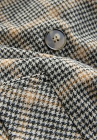 Wollstoff mit einem schwarz-weißen und beige Hahnentrittmuster, ausgestattet mit einem dunklen Vierlochknopf und sichtbaren Nahtnähten.
