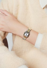Montre en métal argenté et doré avec un cadran rectangulaire, un fond blanc, des index noirs, et un bracelet à maillons bicolore. Portée au poignet.