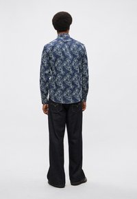 Μπλε ναυτικού χρώματος πουκάμισο με μακριά μανίκια και περίτεχνο ανοιχτό μπλε λουλουδάτο σχέδιο, με μανσέτες με κουμπιά, φορεμένο με σκούρο τζιν παντελόνι με καμπάνα στο κάτω μέρος.