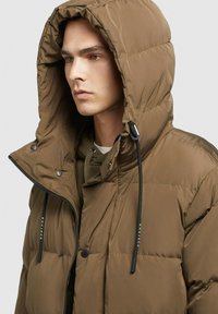 Pruunist nailonist puffer-jakk, millel on pehme tekstuur, suur kapuuts, reguleeritavad paelad, tõmblukk ja nööbid, ning logodetail.