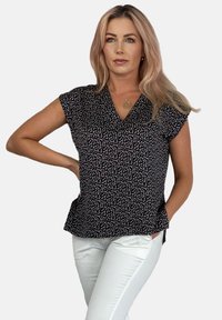 Czarna bluzka z krótkim rękawem, z dekoltem w kształcie litery V i wzorem w białe kropki. Materiał ma miękką teksturę. Połączona z jasnymi spodniami.