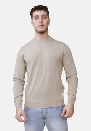 Giovane uomo con capelli corti che indossa un maglione beige a maniche lunghe e jeans azzurri, in piedi davanti a uno sfondo chiaro e uniforme.